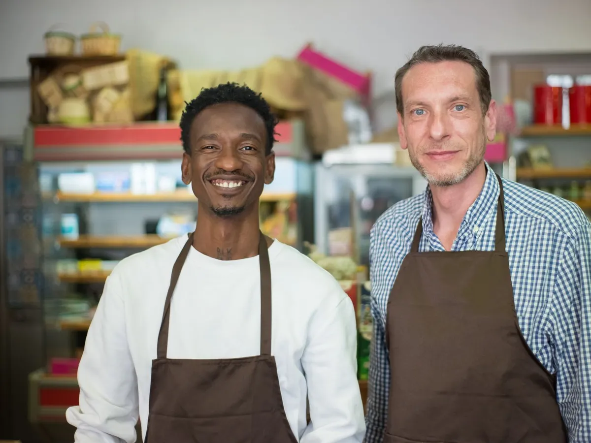 Dois donos de um pequeno comércio, de avental, sorrindo lado a lado atrás do balcão