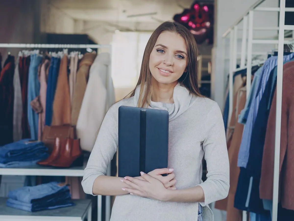 Dona de uma loja de roupas segurando um tablet, sorrindo entre as araras de roupa